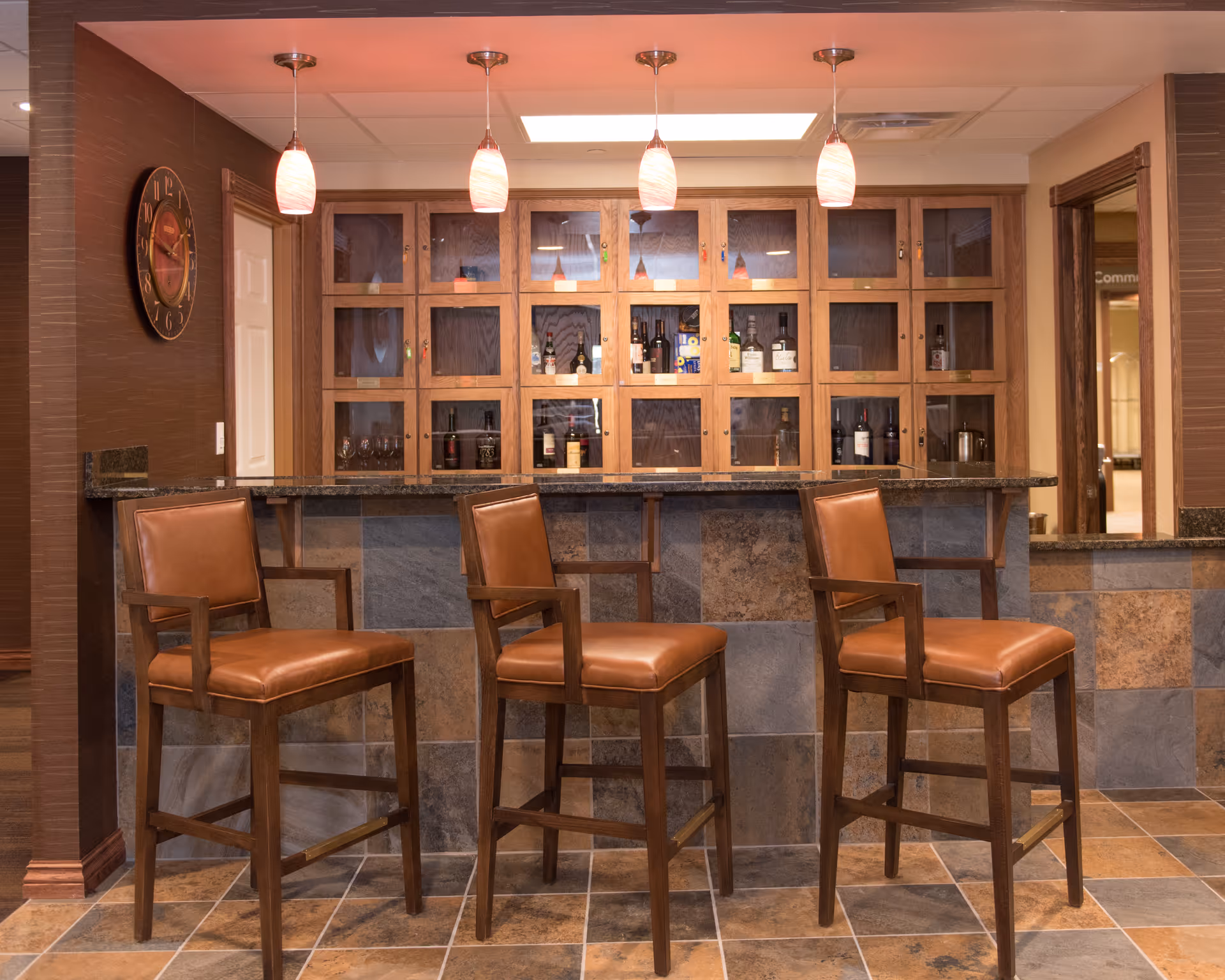 A cozy indoor bar area with three leather bar stools facing a granite countertop and wooden cabinets stocked with bottles.