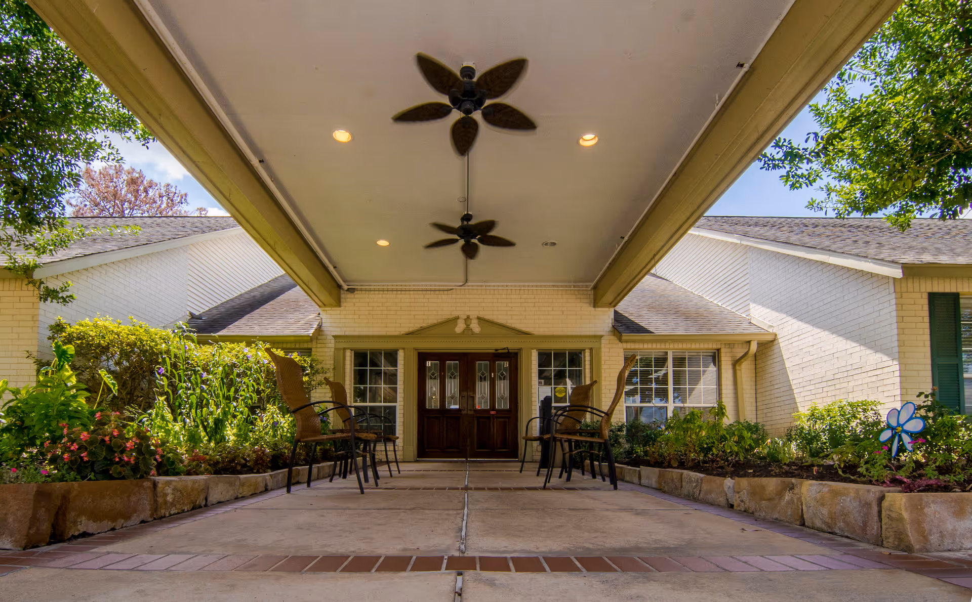 Covered entrance area of a building with two ceiling fans and recessed lighting. There are chairs arranged on both sides along the walkway, with flower beds and greenery bordering the path. The building has light-colored brick walls and a double wooden door with glass panels in the center.