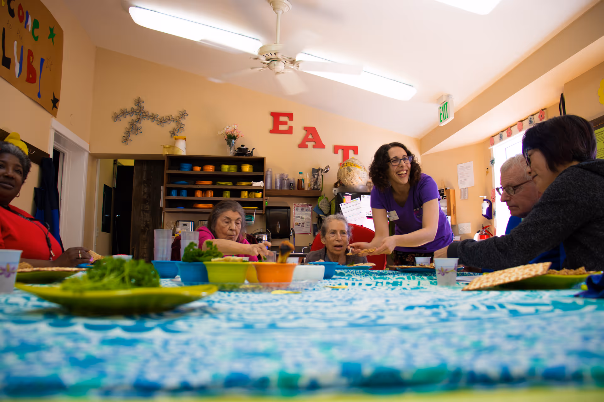 A group of elderly people and a caregiver sitting around a table with food in a brightly lit room. The caregiver is smiling and interacting with the group. The wall behind them has shelves with colorful dishes and large red letters spelling 'EAT'.