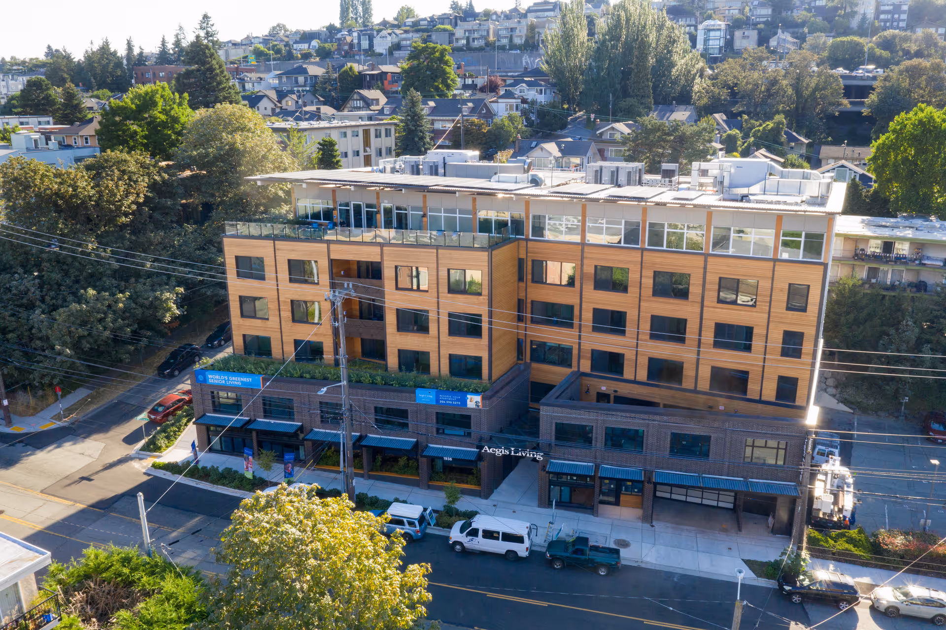 Aerial view of a multi-story senior living facility named Aegis Living Lake Union, featuring a modern building with large windows, a rooftop terrace, and surrounded by trees and residential houses.