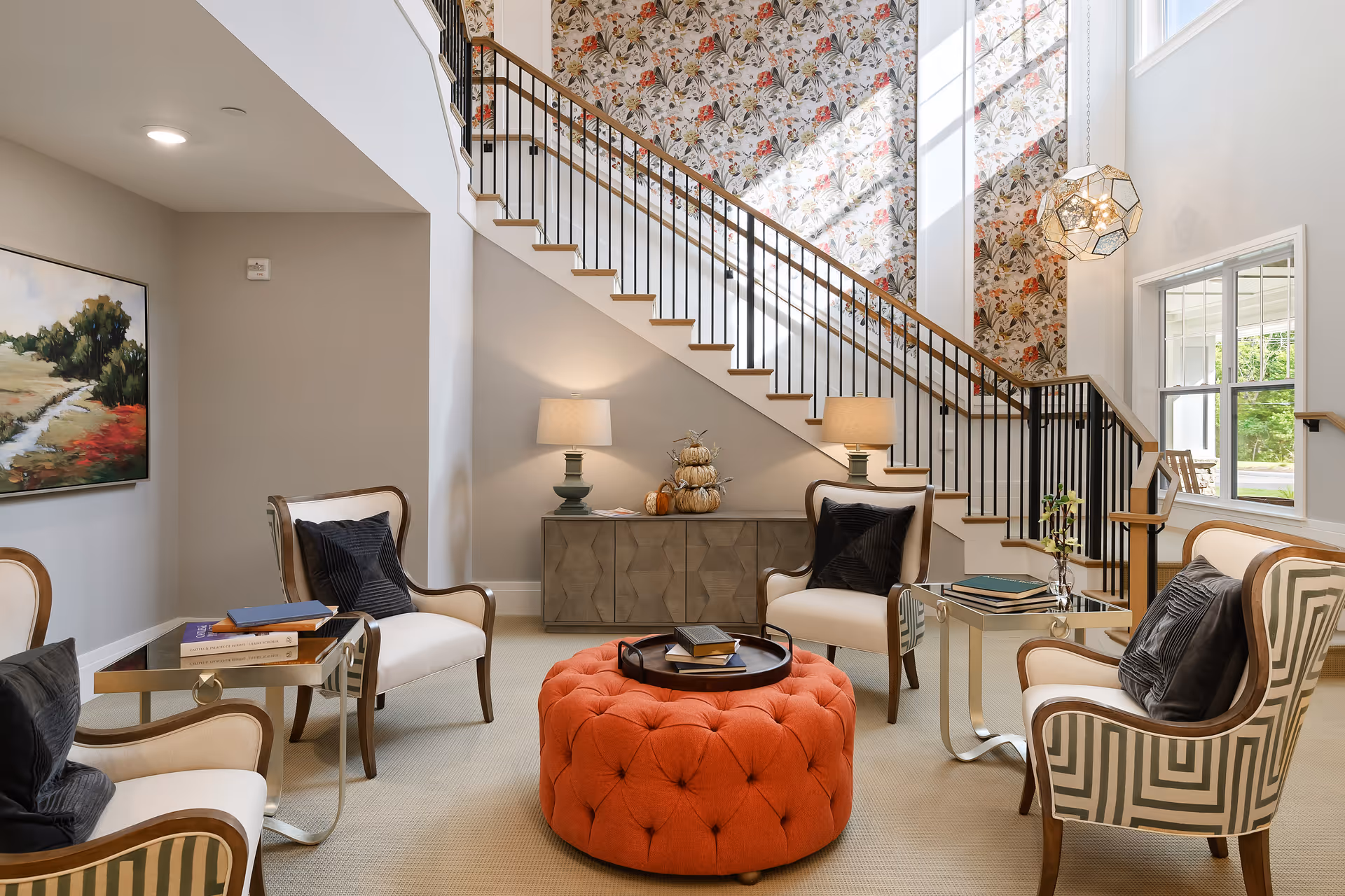 A bright and inviting living room area featuring four upholstered armchairs with dark cushions arranged around a large, round, tufted orange ottoman. There are two small side tables with books and decorative items. Behind the seating area is a staircase with a black metal railing and floral wallpaper on the wall behind it. Two table lamps sit on a console table under the stairs, and a large window lets in natural light.