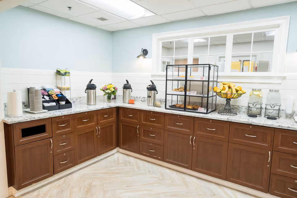 A self-serve refreshment counter with coffee dispensers, pastries, fruit, and water stations on a marble countertop in a communal area.