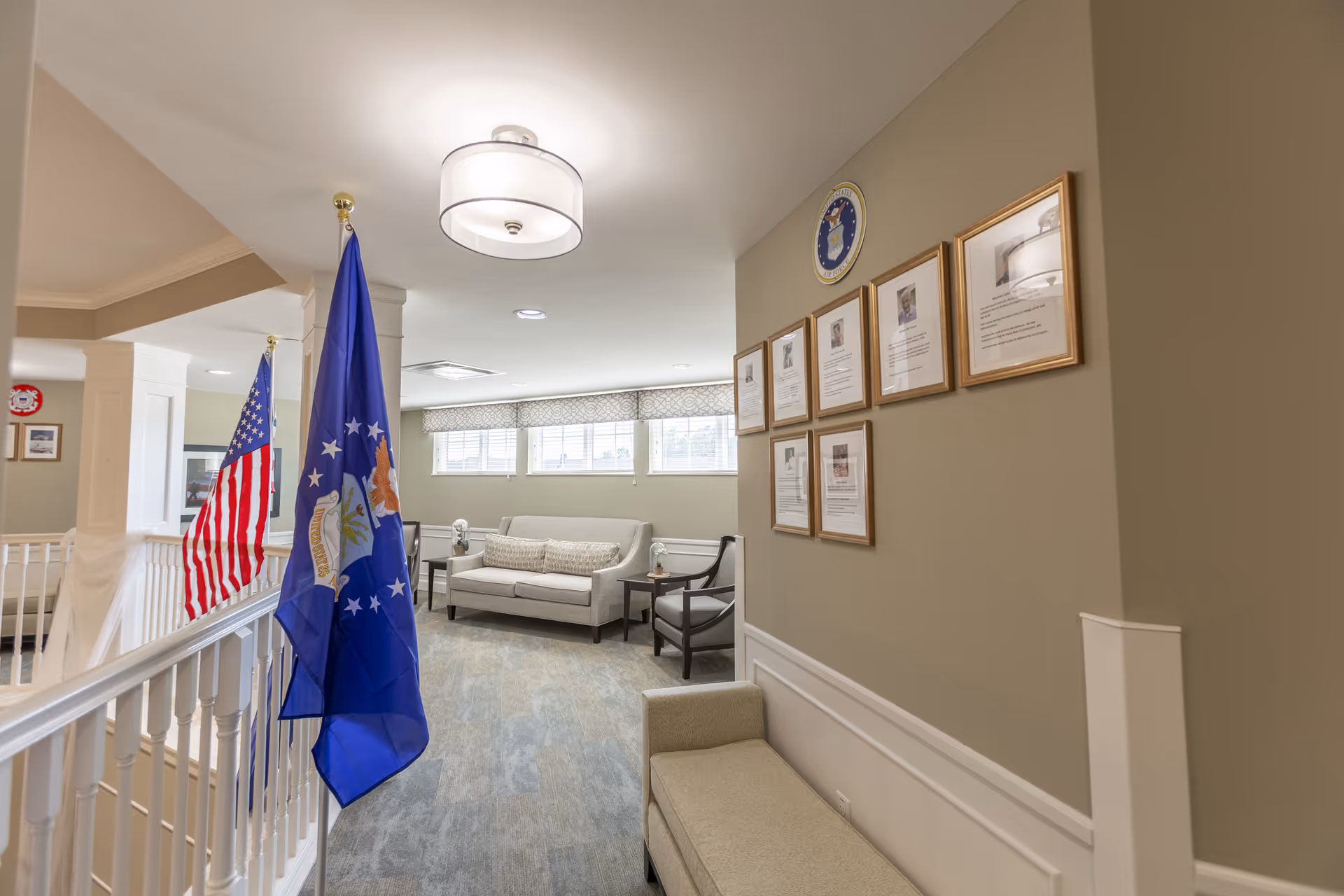 Hallway lounge in a senior living facility with a sofa and chairs, framed photos on the wall, and U.S. and blue flags displayed.