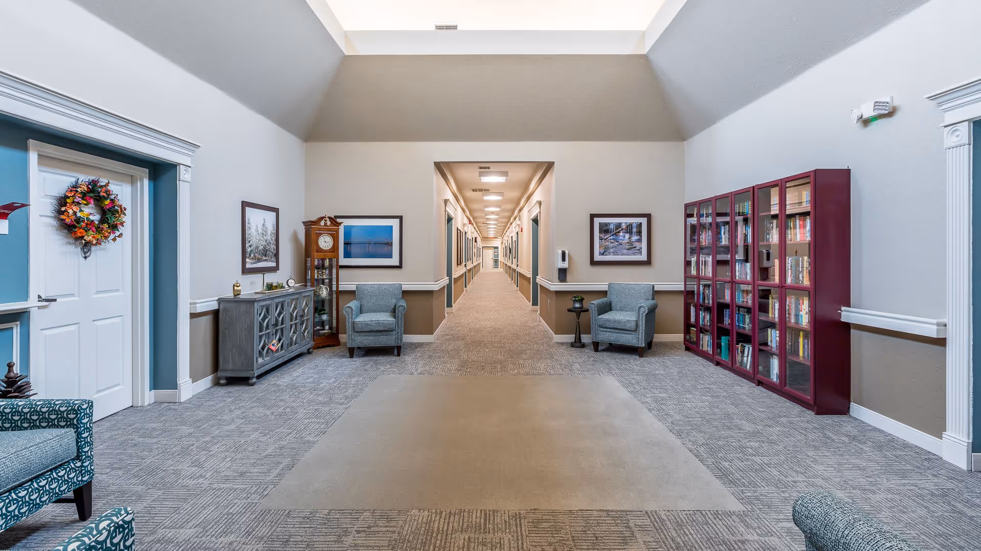 A spacious hallway in an assisted living facility with a high ceiling and skylight. The hallway is carpeted and lined with framed pictures on the walls. There are two blue armchairs with a small table between them on the right side, a tall grandfather clock, and a decorative cabinet on the left side. A large bookshelf filled with books is also visible on the right. The hallway extends into the distance with multiple doors along the sides.