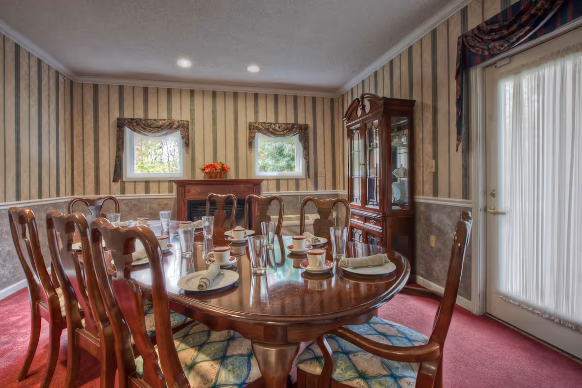 A formal dining room with a polished wooden dining table set for eight, featuring cups, plates, napkins, and glasses. The room has striped wallpaper, two small windows with floral valances, a wooden china cabinet, and a door with sheer curtains. The floor is carpeted in red.