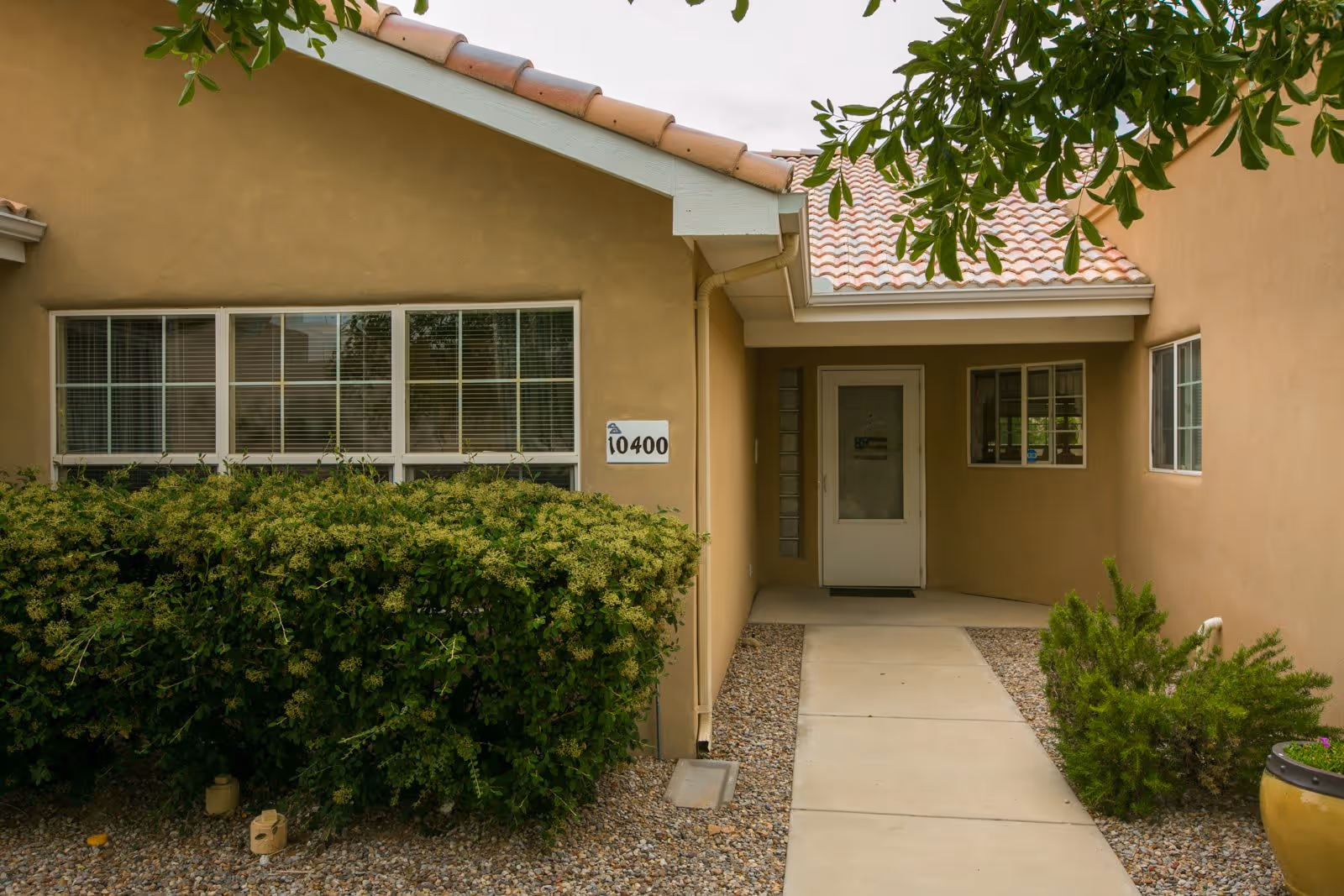 Entrance to a building with a beige exterior, a tiled roof, and a concrete walkway leading to a white door. There are green bushes and plants on either side of the walkway, and windows with white frames and blinds are visible on the walls.