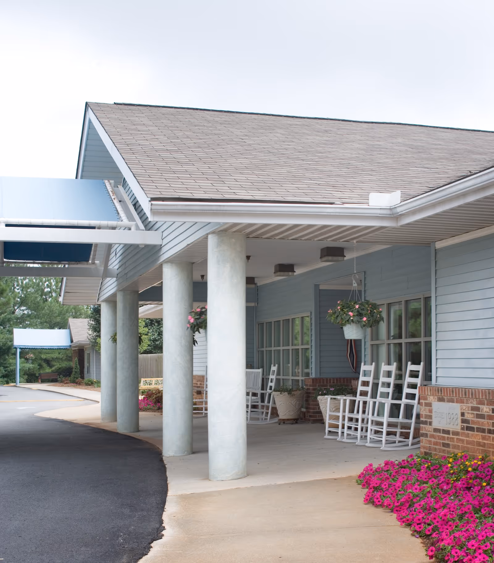 Covered entrance area of a building with white columns, light blue siding, and a row of white rocking chairs on the porch. There are hanging flower pots and bright pink flowers planted near the walkway.