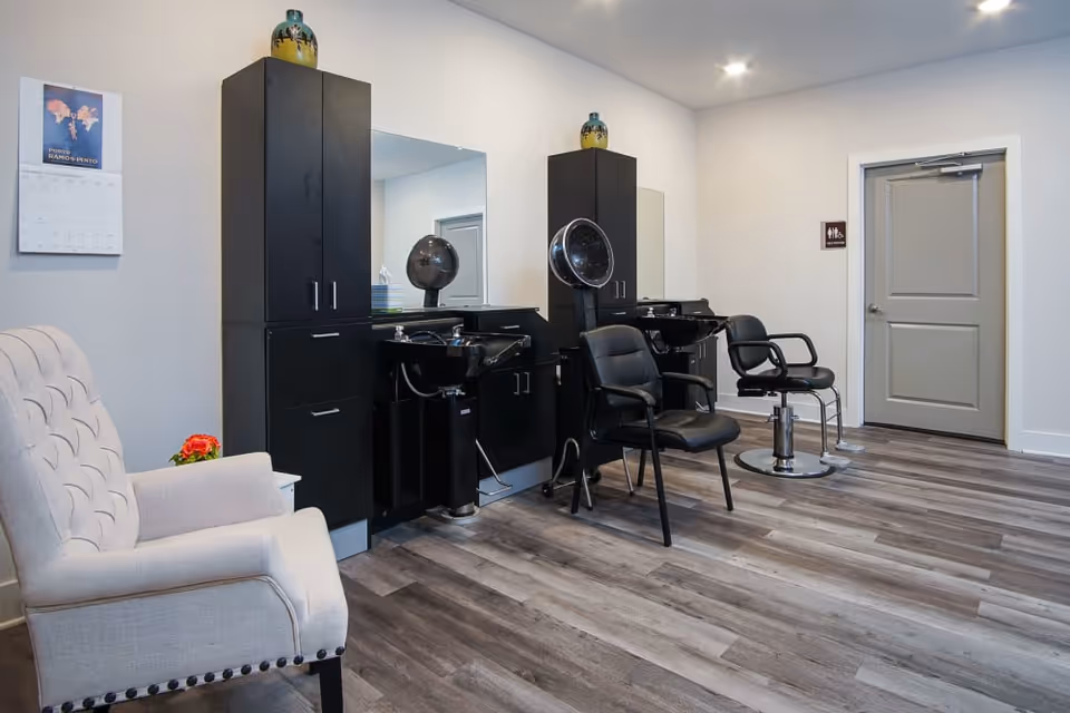 Interior view of a salon area in a senior living facility with two black salon chairs, hair washing sinks, tall black cabinets, a beige upholstered armchair, a calendar on the wall, and a closed gray door.