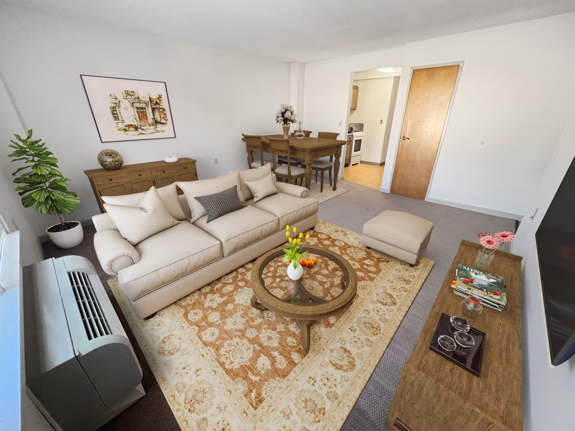 A cozy living room with a beige sofa, matching ottoman, and a round wooden coffee table with a vase of yellow flowers and a bowl of fruit. There is a patterned area rug under the furniture. Against the wall is a wooden sideboard with decorative items and a framed artwork above it. A dining table with four chairs is visible in the background near an open doorway leading to a kitchen. A large flat-screen TV is mounted on the wall opposite the sofa, and a potted plant is placed near the window.