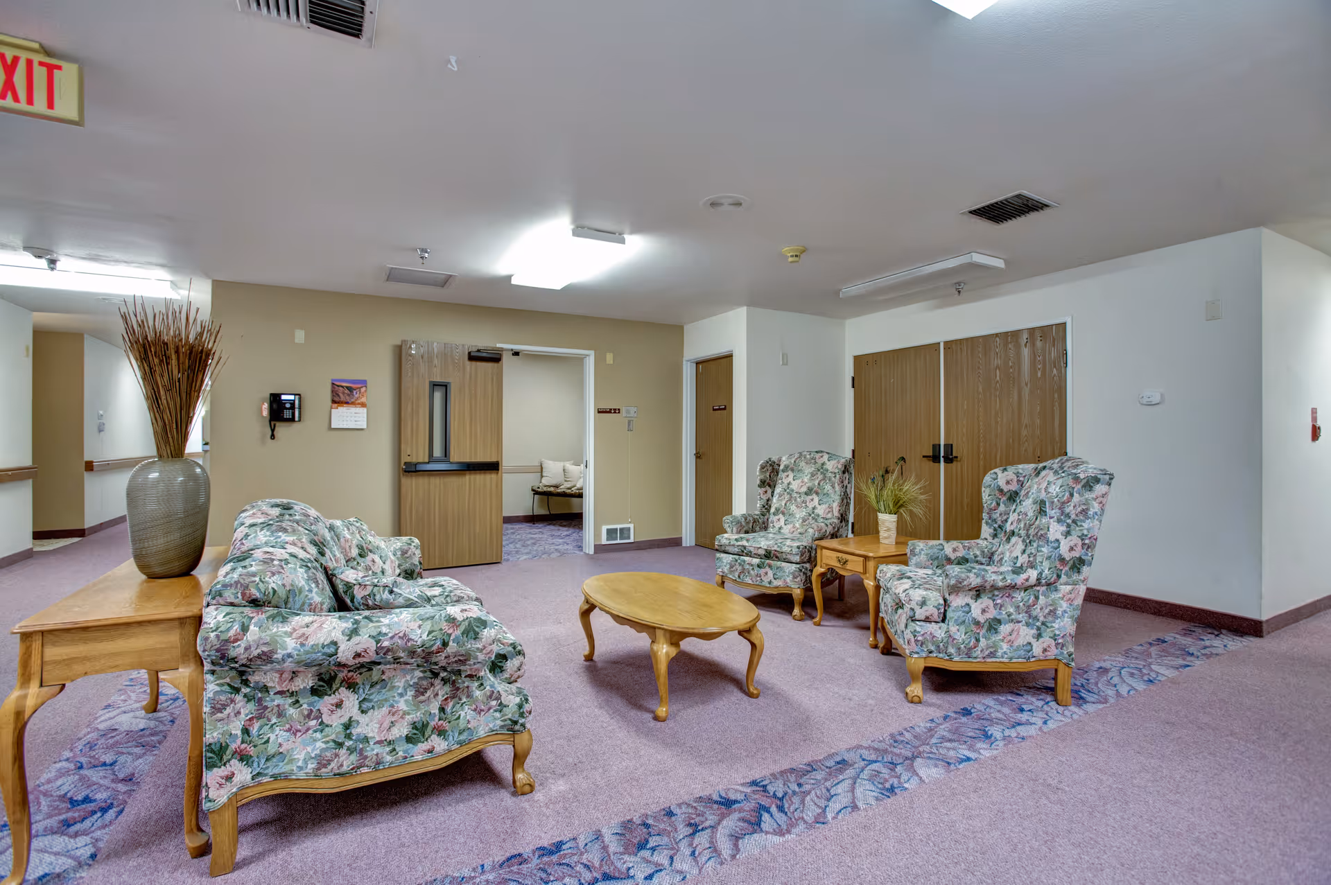 A sitting area in a senior living facility with floral upholstered sofa and two armchairs arranged around a wooden coffee table. There is a wooden side table with a large vase holding decorative branches. The room has beige walls, purple carpet with a floral border, and several wooden doors. An exit sign is visible on the ceiling.