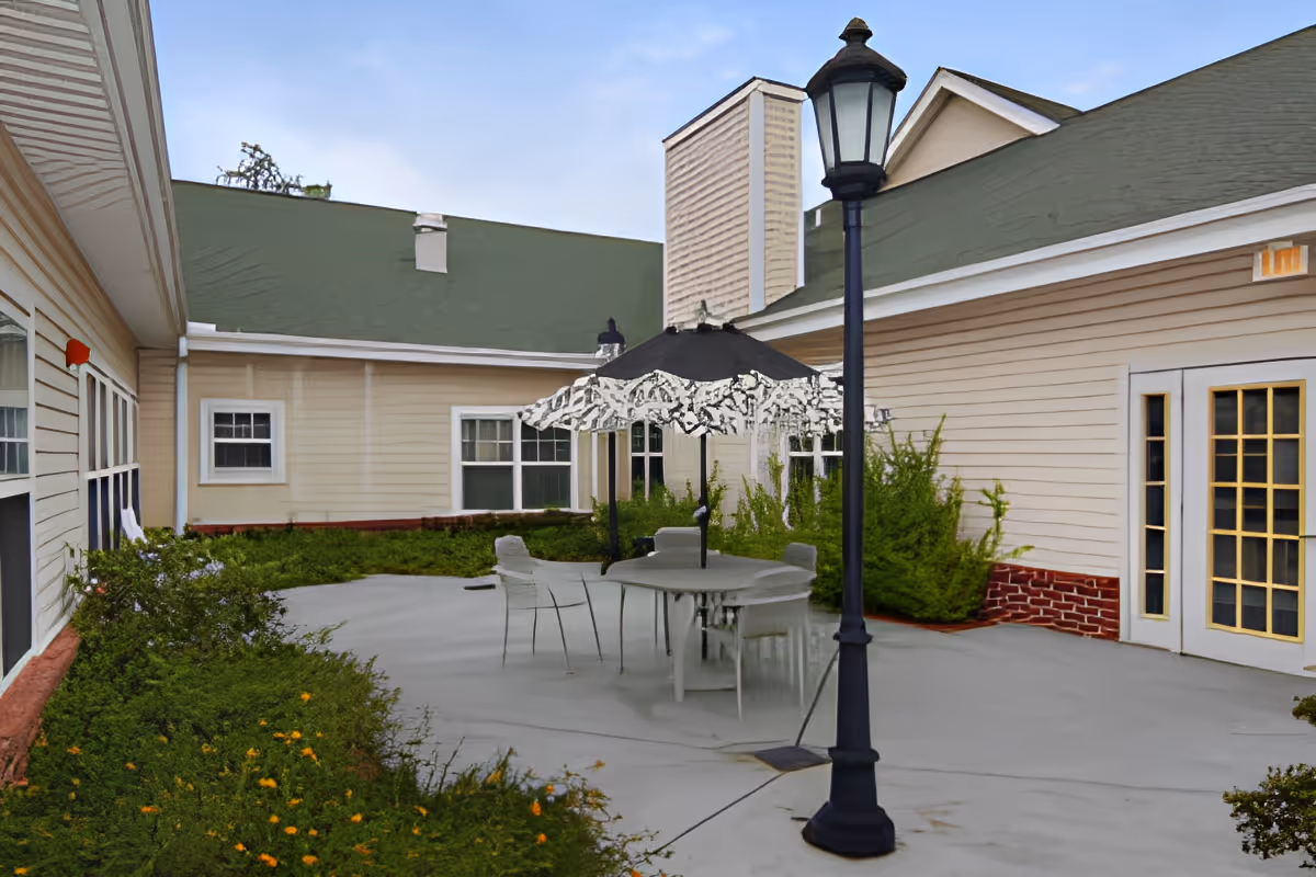 Outdoor patio area at TerraBella Southern Pines featuring a round table with four chairs and a large umbrella, surrounded by beige buildings with green roofs and shrubbery along the edges. A black lamp post stands near the table on the concrete patio.