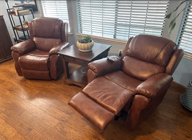 Two brown leather recliner chairs positioned on a wooden floor next to a wooden side table with a potted plant on top, in front of a window with white blinds.