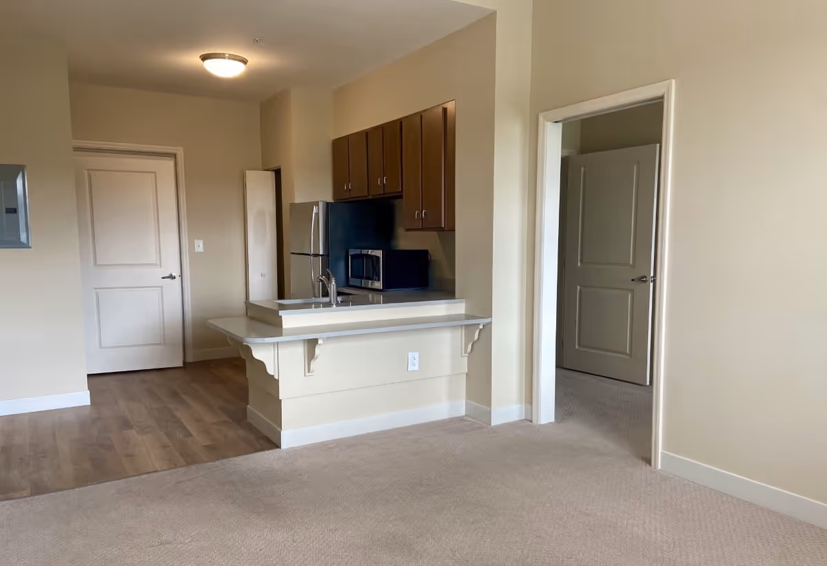 Interior view of a senior living facility apartment showing a small kitchen area with wooden cabinets, a refrigerator, a microwave, and a sink with a countertop. The floor transitions from wood in the kitchen to carpet in the adjacent room. There are two open doorways leading to other rooms.