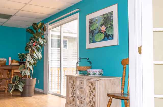 Interior view of a room with turquoise walls featuring a wooden sideboard with decorative items on top, a wooden chair, a large potted plant, a framed painting of water lilies on the wall, and a sliding glass door leading outside.