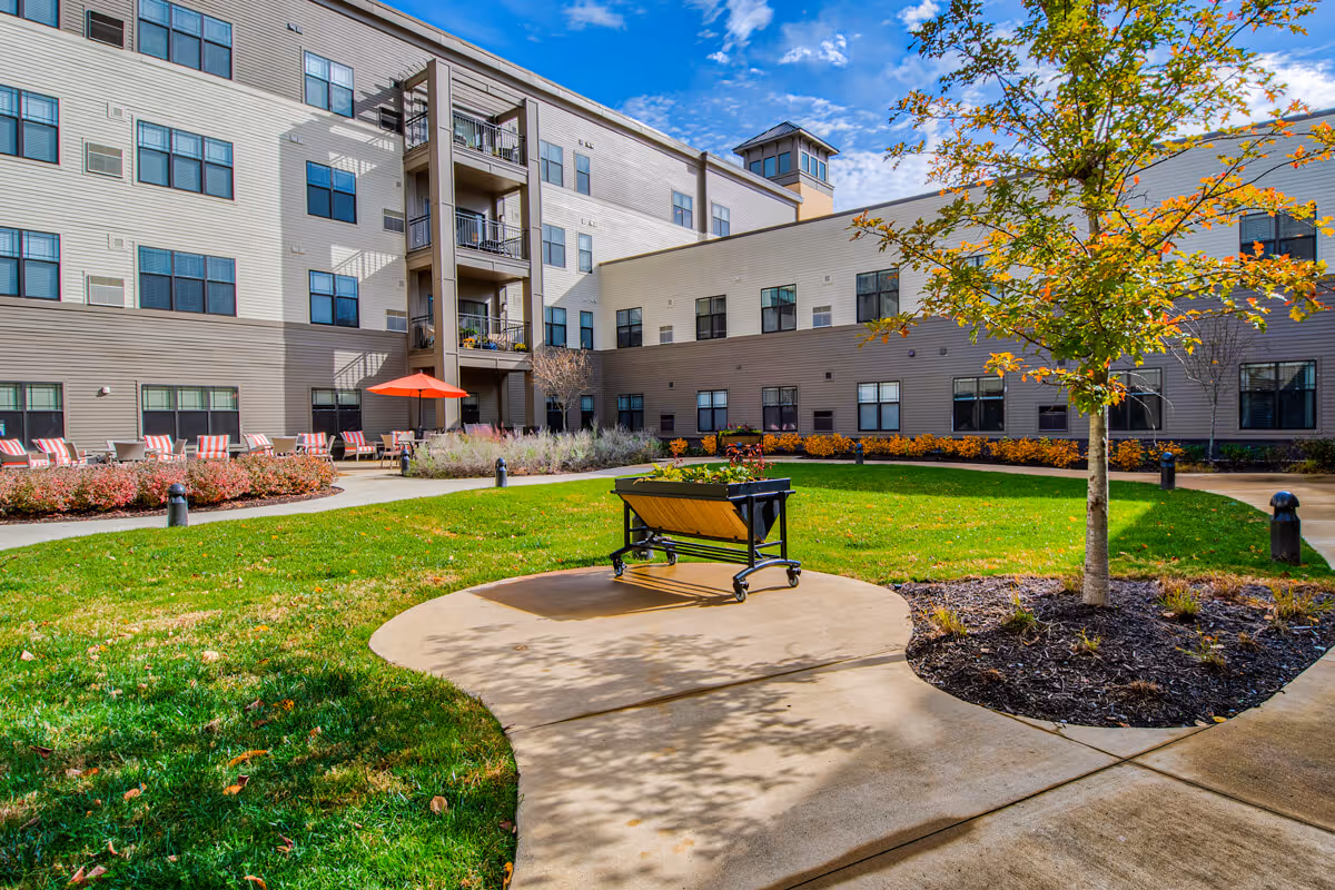 Outdoor courtyard area of a senior living facility with green grass, a small tree, a raised garden bed on wheels, and a paved walkway. The multi-story building with numerous windows surrounds the courtyard, and there are several chairs with red and white striped cushions and a red umbrella near the building. The sky is blue with some clouds.