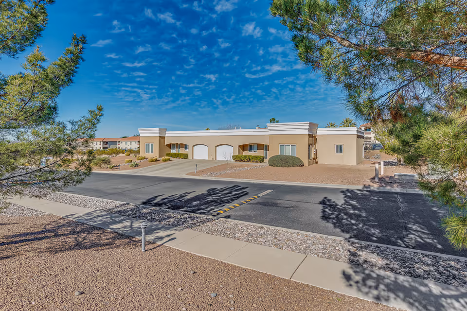 Exterior view of a single-story beige building with white trim, featuring two garage doors and several windows, surrounded by desert landscaping with gravel and bushes. The scene is framed by green pine trees under a blue sky with scattered clouds.