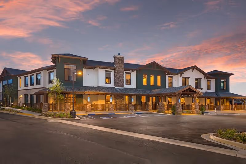 Exterior view of a two-story senior living facility building at dusk with warm interior lights visible through the windows, a stone chimney, covered entrance, and a parking area with handicap spaces in front.