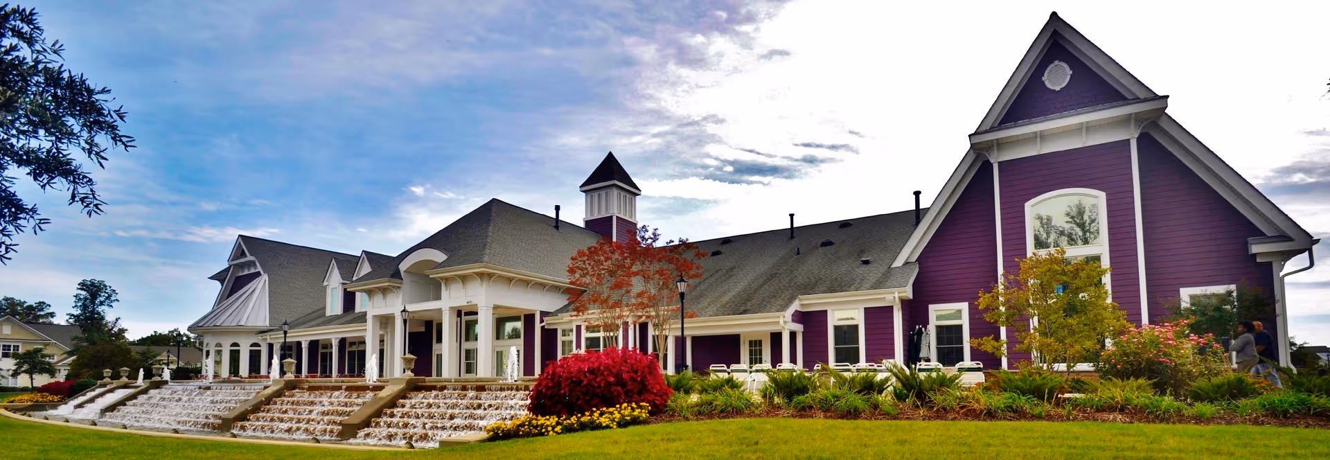 Exterior view of The Willows at Victoria Falls, a large building with purple siding and white trim, featuring a multi-level cascading water fountain in front, landscaped gardens with colorful flowers and shrubs, and a partly cloudy sky overhead.