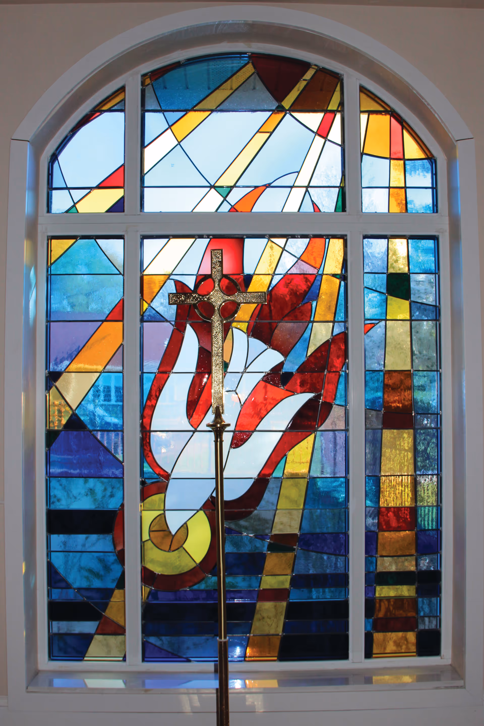 A colorful stained glass window featuring abstract shapes in red, blue, yellow, and white. In front of the window stands a gold-colored cross on a tall stand.