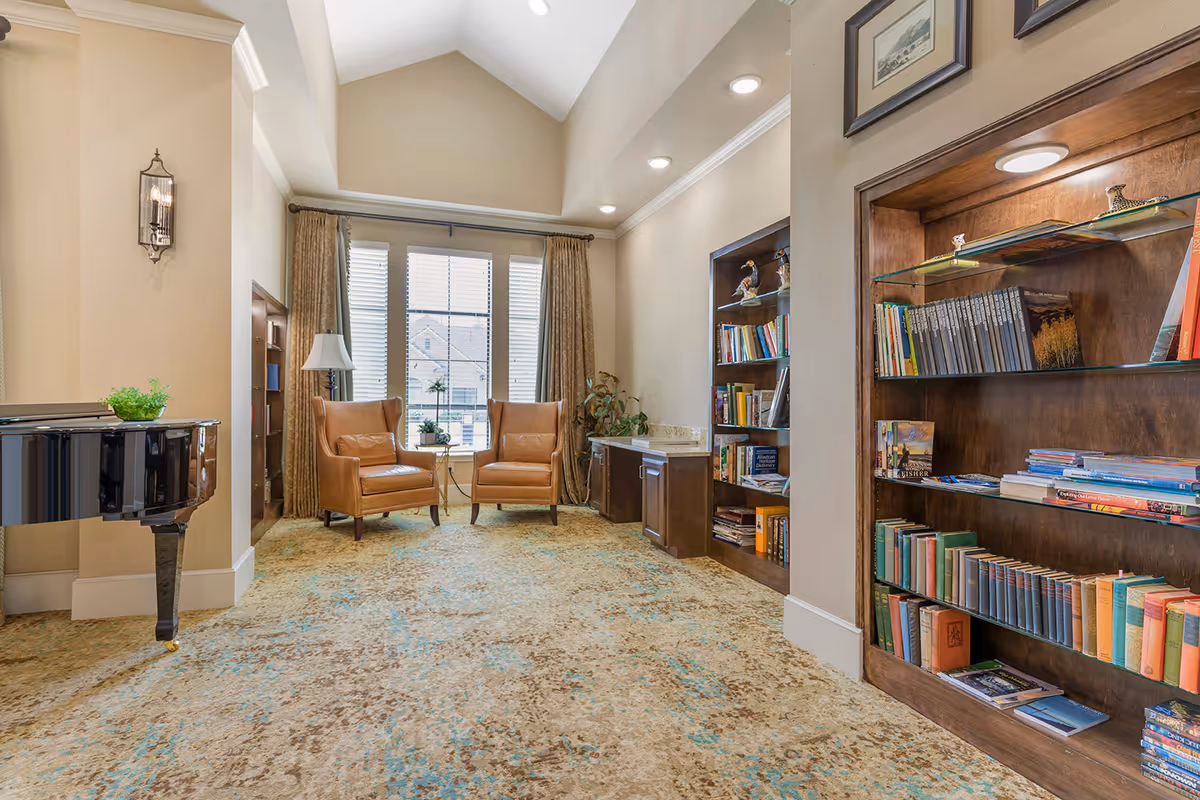 A cozy senior living common area with two brown leather armchairs facing a large window with beige curtains. There is a black grand piano on the left and built-in wooden bookshelves filled with books on the right. The room has a high vaulted ceiling with recessed lighting and a patterned carpet in shades of beige and blue.