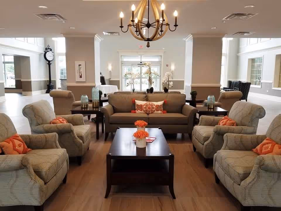 A spacious and well-lit living room area with a central dark wooden coffee table decorated with orange flowers. Surrounding the table are four beige armchairs with orange patterned pillows and a brown sofa with a decorative pillow. The room features large windows, a chandelier, and light-colored walls with white trim.
