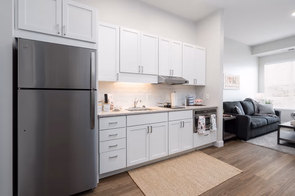 A modern kitchen with white cabinets, a stainless steel refrigerator, a sink, stovetop, and oven. The kitchen opens into a living room area with a dark gray sofa, a coffee table, and a window with blinds letting in natural light.