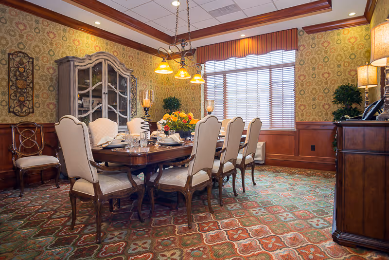 A traditional dining room with a large wooden dining table surrounded by eight upholstered chairs. The table is set with glassware, napkins, and a floral centerpiece. The room features patterned wallpaper, a decorative carpet, a wooden cabinet with glass doors, and a window with blinds and a valance. Warm lighting is provided by a chandelier and table lamps.
