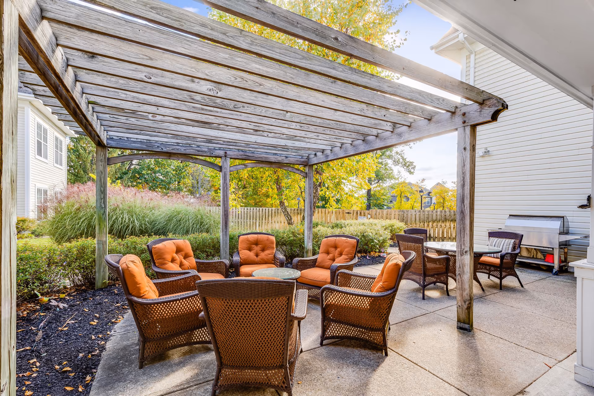Outdoor patio area at Sunrise of Morris Plains featuring a wooden pergola overhead, cushioned wicker chairs arranged around a small round table, additional seating with a glass-top table, and a barbecue grill. The area is surrounded by greenery and a wooden fence with trees showing autumn foliage in the background.