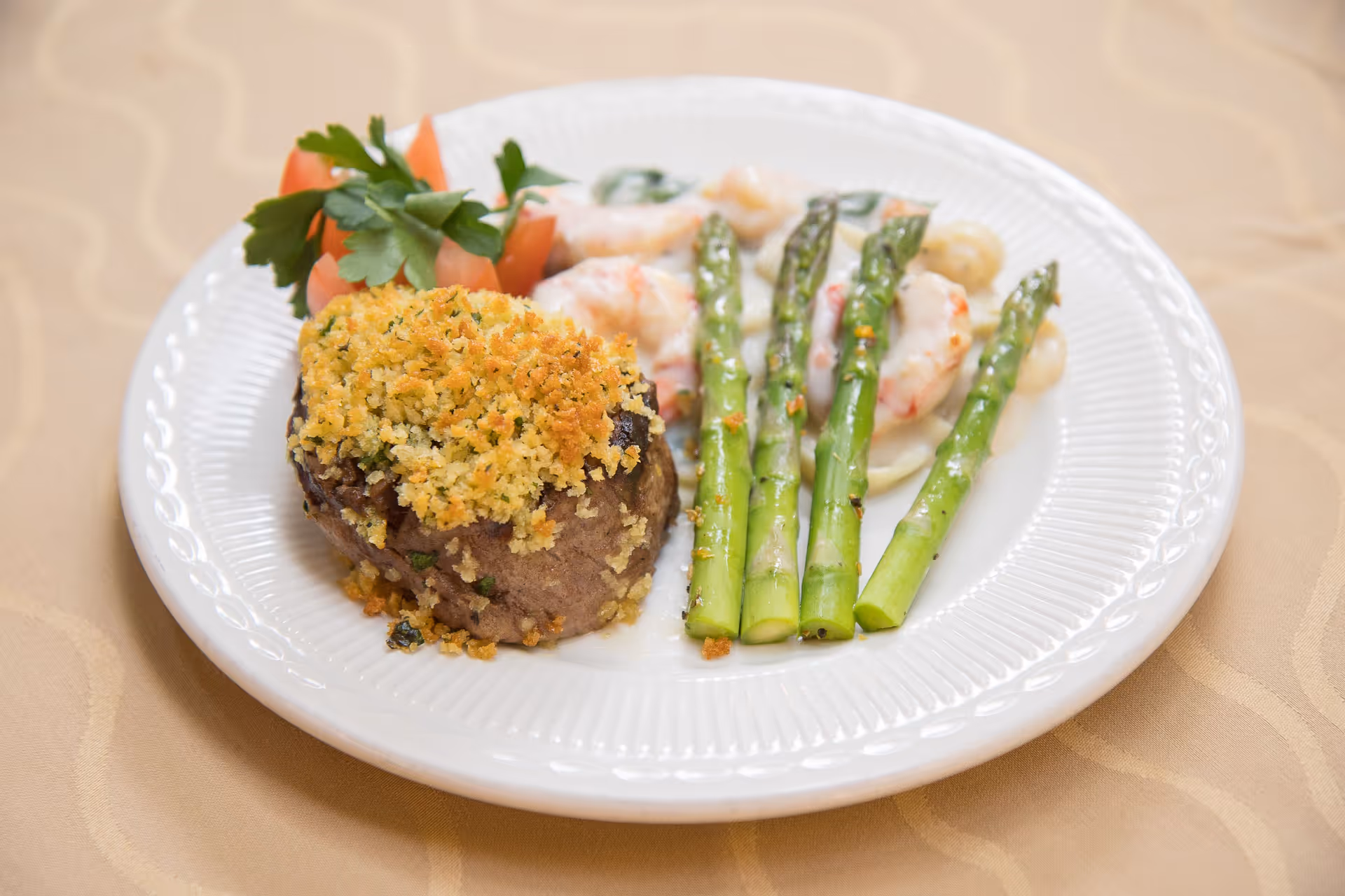 A white plate with a serving of breaded and baked meat topped with golden breadcrumbs, steamed asparagus spears, shrimp in a creamy white sauce, and a garnish of tomato slices and parsley on a beige tablecloth.