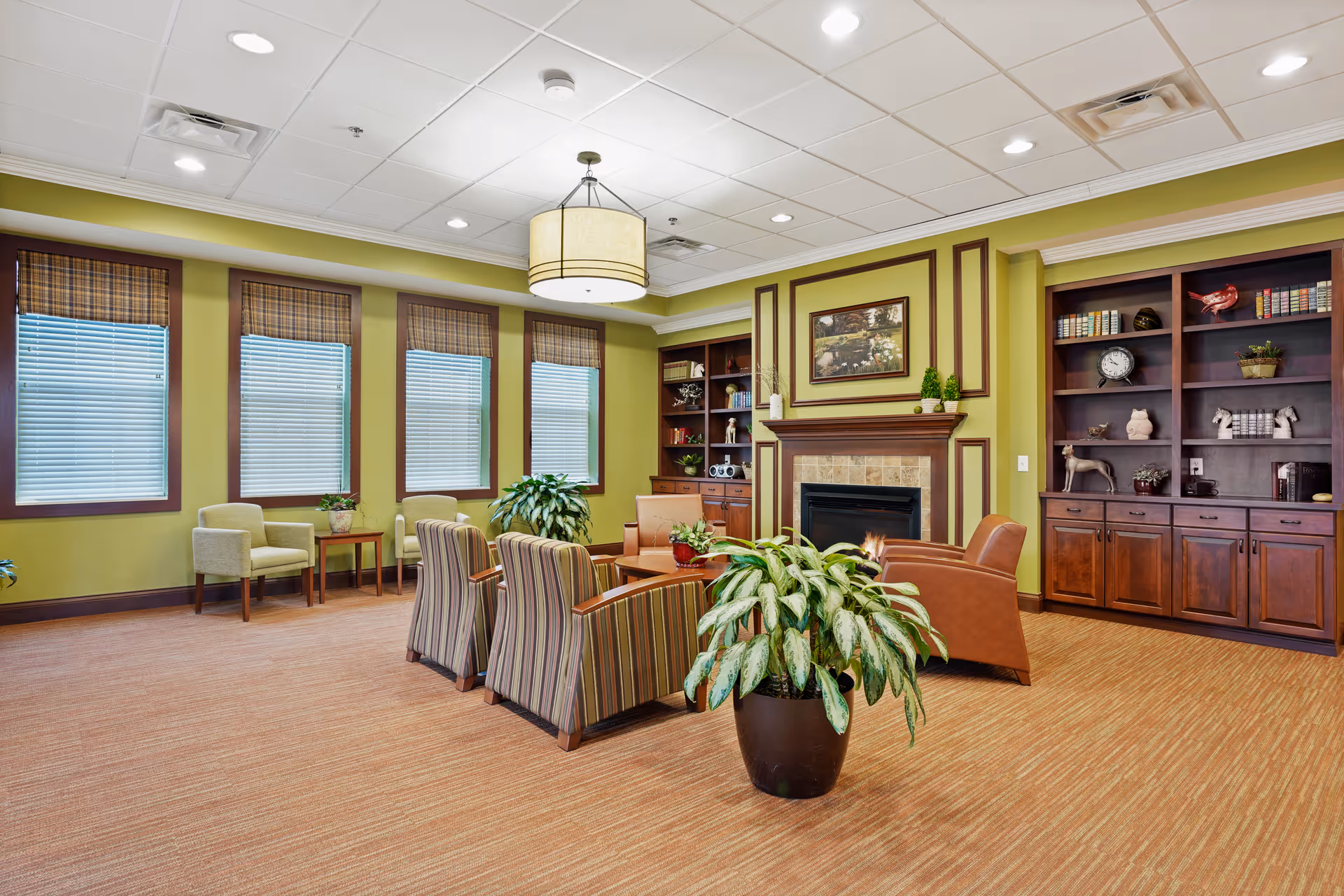 A spacious and well-lit living room area with green walls and large windows covered with blinds. The room features a fireplace with a framed painting above it, flanked by built-in wooden bookshelves filled with decorative items and books. There are several comfortable chairs arranged around a coffee table, and a large potted plant is placed in the foreground. The ceiling has recessed lighting and a central hanging light fixture.