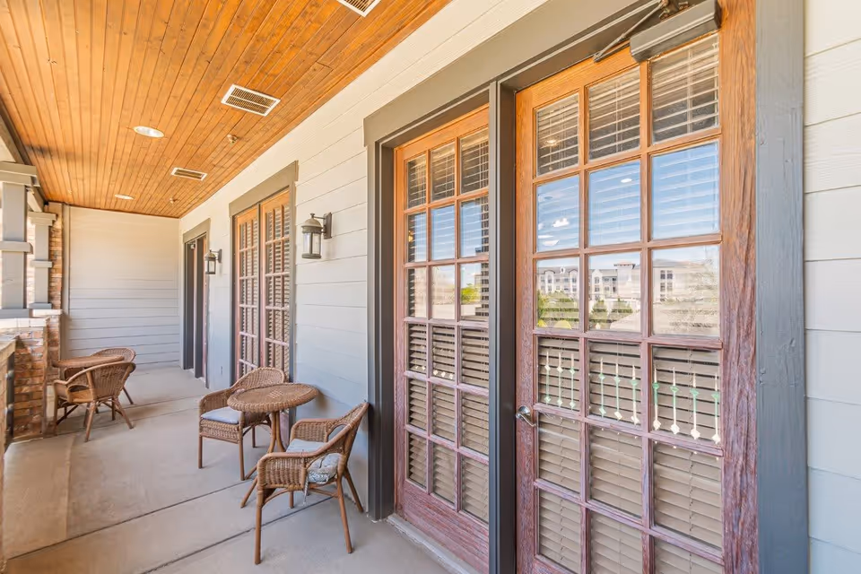 A covered outdoor balcony area with a wooden ceiling, beige walls, and multiple sets of wooden-framed glass doors with blinds. The balcony has wicker chairs and small round tables arranged for seating.