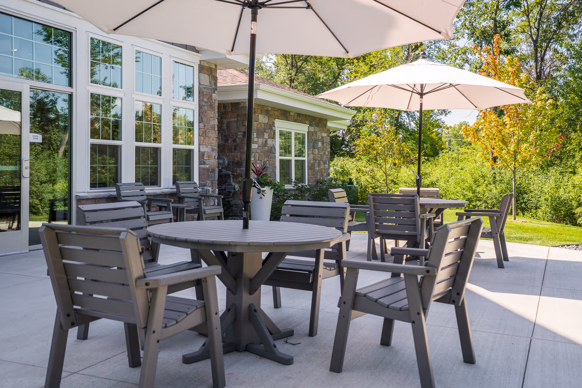 Outdoor patio area at NorBella Senior Living - Champlin featuring round tables with umbrellas and multiple gray chairs on a concrete surface, surrounded by greenery and trees with a stone building in the background.
