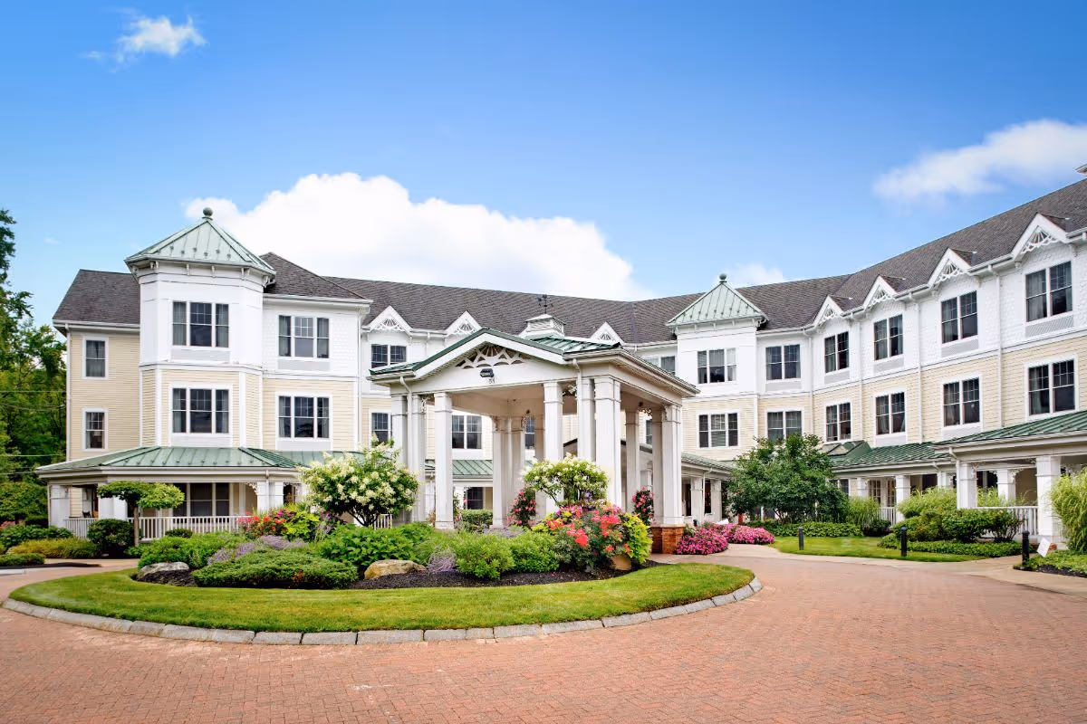 Front exterior of a large three-story senior living building with a covered porte-cochère, landscaped circular driveway, and colorful flower beds.