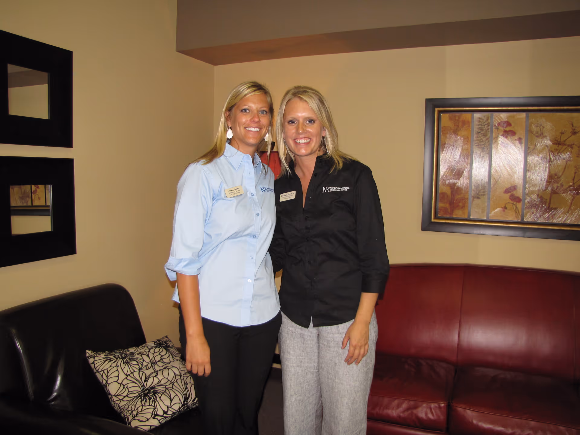 Two women standing and smiling in a living room area with a black leather chair, a red leather couch, decorative pillows, framed artwork on the wall, and beige walls.