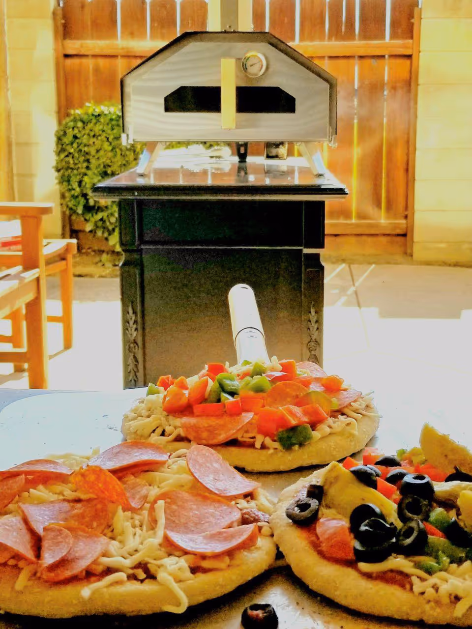 Three uncooked personal pizzas topped with pepperoni, bell peppers, olives, and cheese on a table outdoors with a pizza oven and wooden fence in the background.