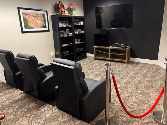 Small media room with three black recliners facing a wall-mounted TV, a bookshelf and a red velvet rope barrier.