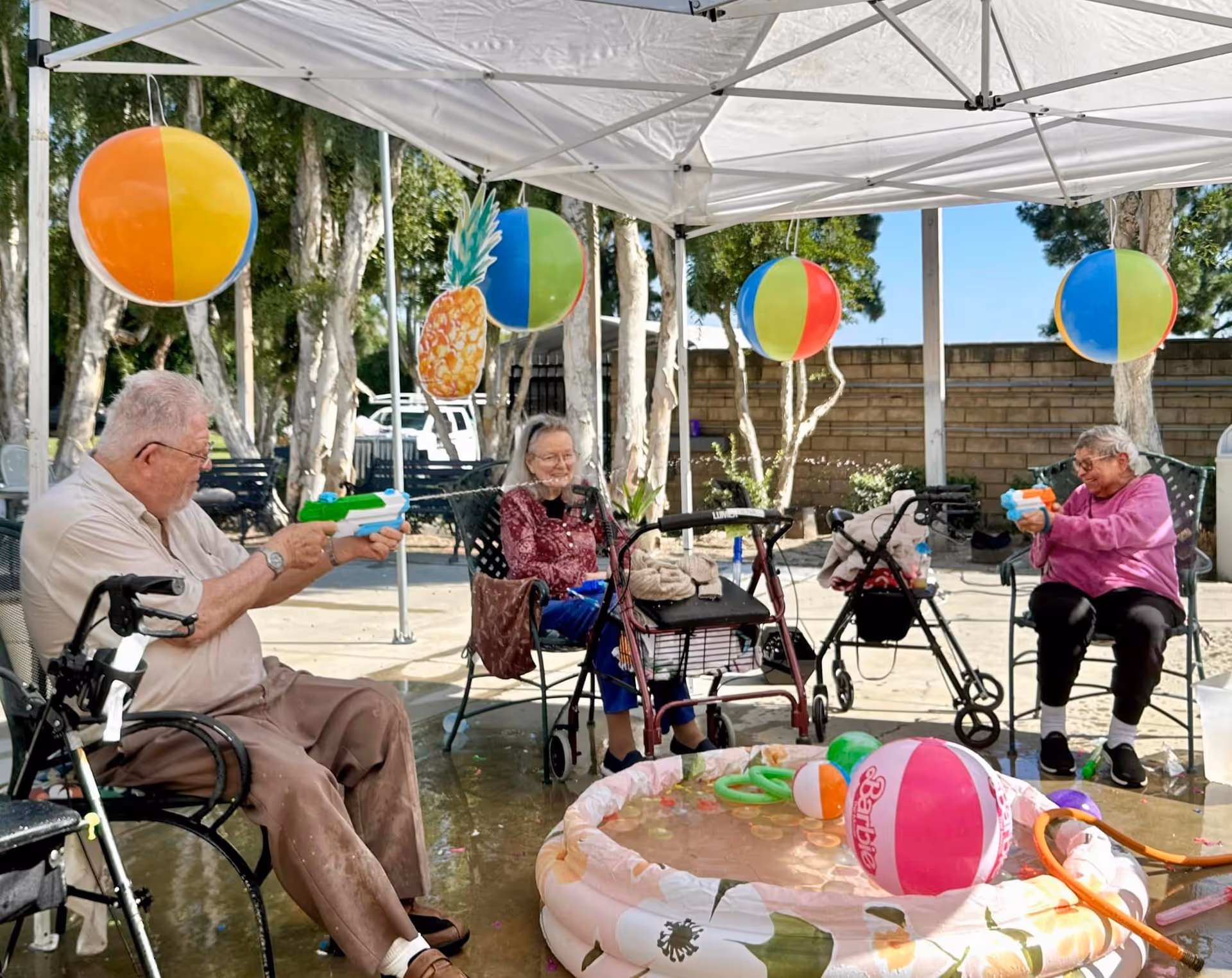 Three elderly individuals sitting outdoors under a canopy, playing with water guns and surrounded by colorful beach balls and a small inflatable pool filled with water. Two walkers are visible near the seated individuals, and trees and a brick wall are in the background.