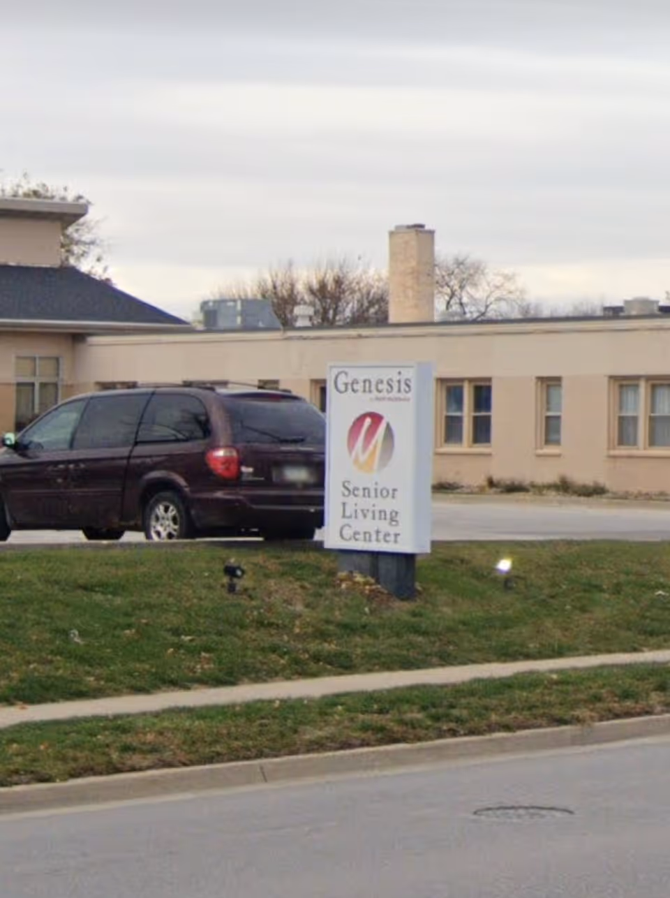 Exterior view of Genesis Senior Living Center showing a beige building with multiple windows, a parked dark-colored minivan, and a sign with the facility's name on a grassy area near the sidewalk.