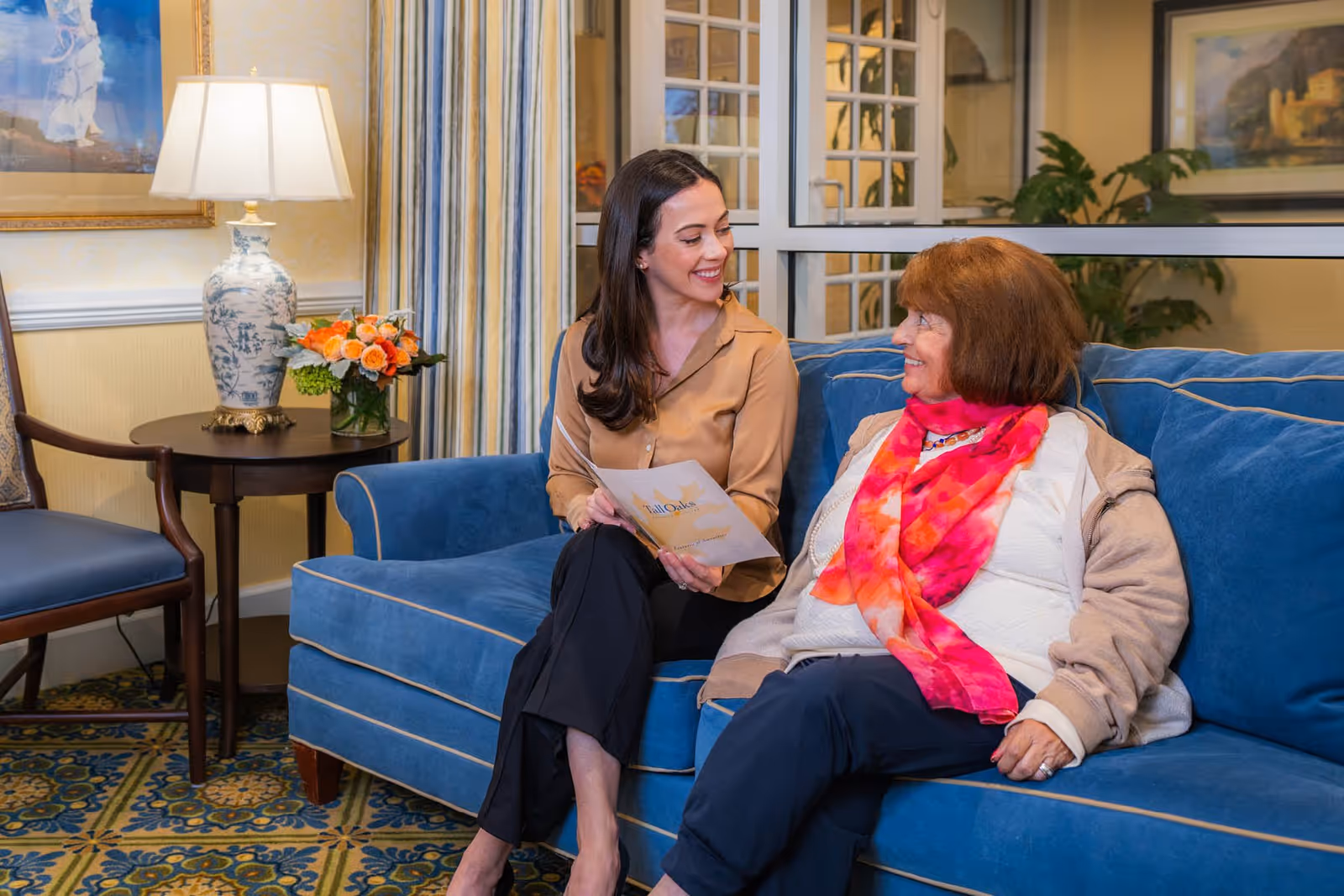 Two women sitting on a blue couch in a cozy living room setting. One woman, younger, is holding a brochure and smiling at the older woman, who is wearing a bright pink and orange scarf. The room features a side table with a decorative lamp and a vase of flowers, patterned carpet, and framed artwork on the walls.
