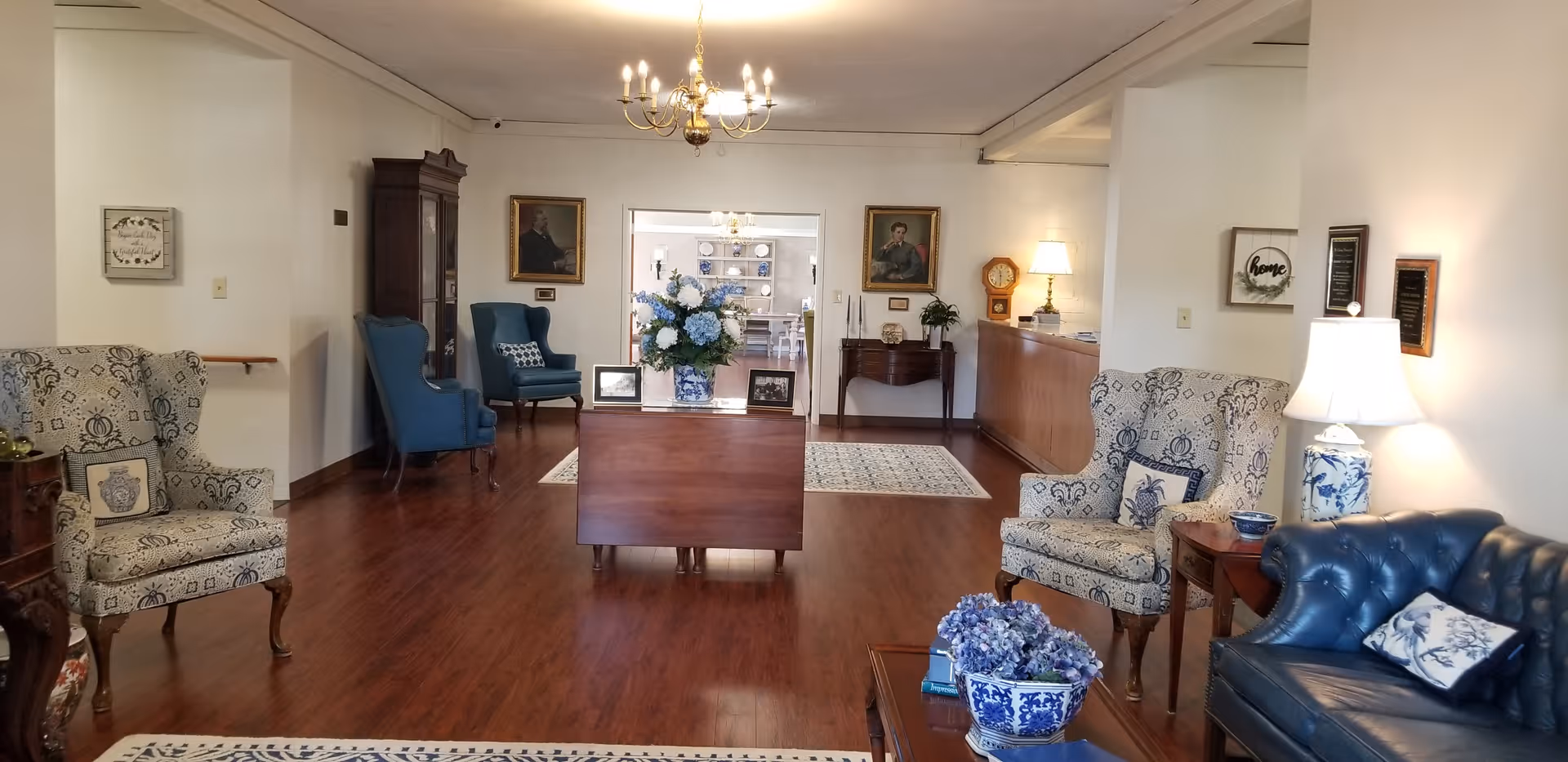 Spacious sitting room with upholstered armchairs, a leather sofa, wooden tables and a central floral arrangement in a senior living facility.