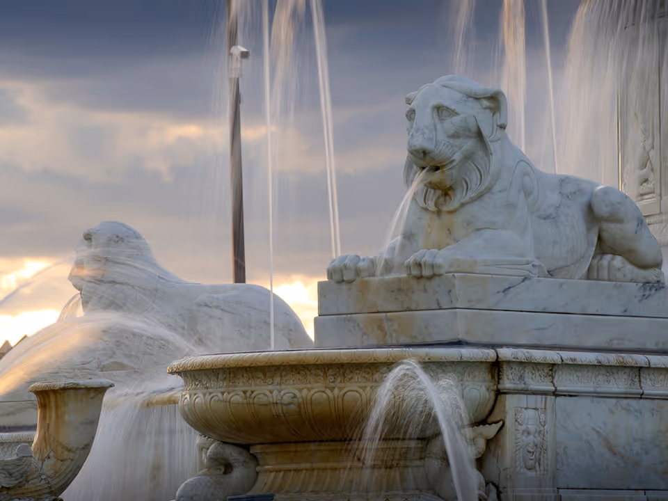 Close-up view of a marble fountain featuring two lion sculptures with water spouting from their mouths, set against a cloudy sky at sunset.