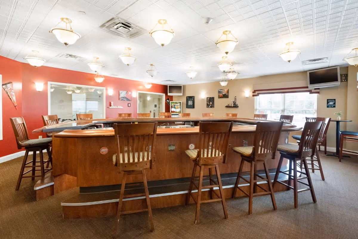 Interior view of a spacious common area with a large wooden bar counter surrounded by high wooden chairs. The room has a red accent wall with framed pictures and a window letting in natural light. Ceiling lights illuminate the space, and there are two wall-mounted televisions in opposite corners.