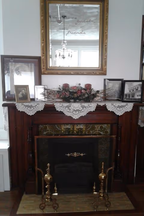 Wooden fireplace mantel topped with a lace runner, framed photos, a floral arrangement, and a mirror reflecting a chandelier.