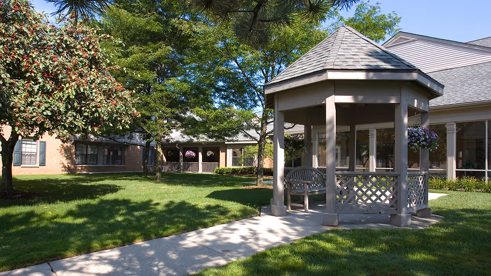 A sunny outdoor courtyard at Oakmont Livonia Senior Community featuring a small gazebo with a bench inside, surrounded by green grass, trees, and a brick building with large windows in the background.