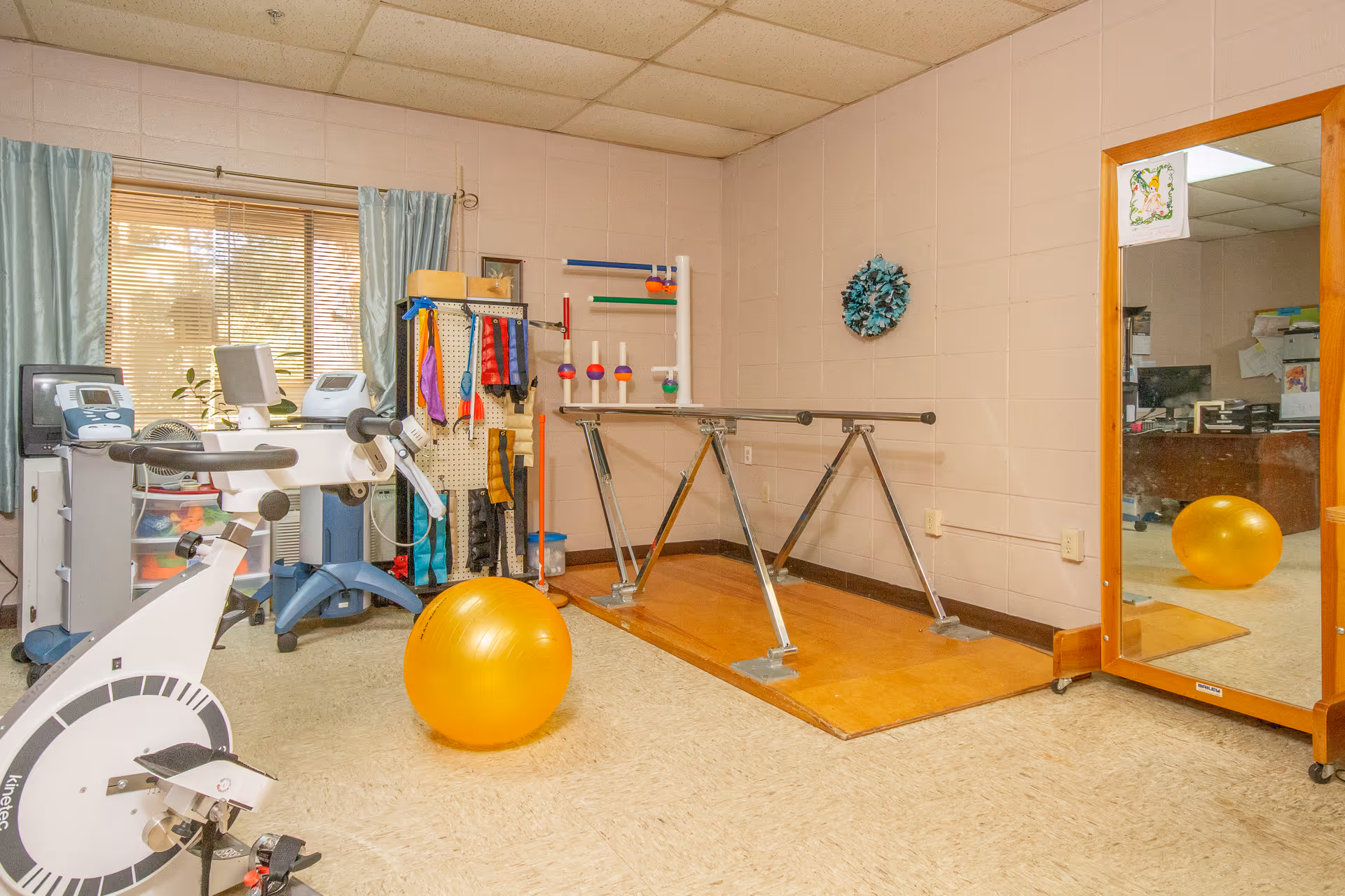A rehab therapy room with parallel bars, an exercise bike, a yellow stability ball, assorted therapy equipment and a large mirror.