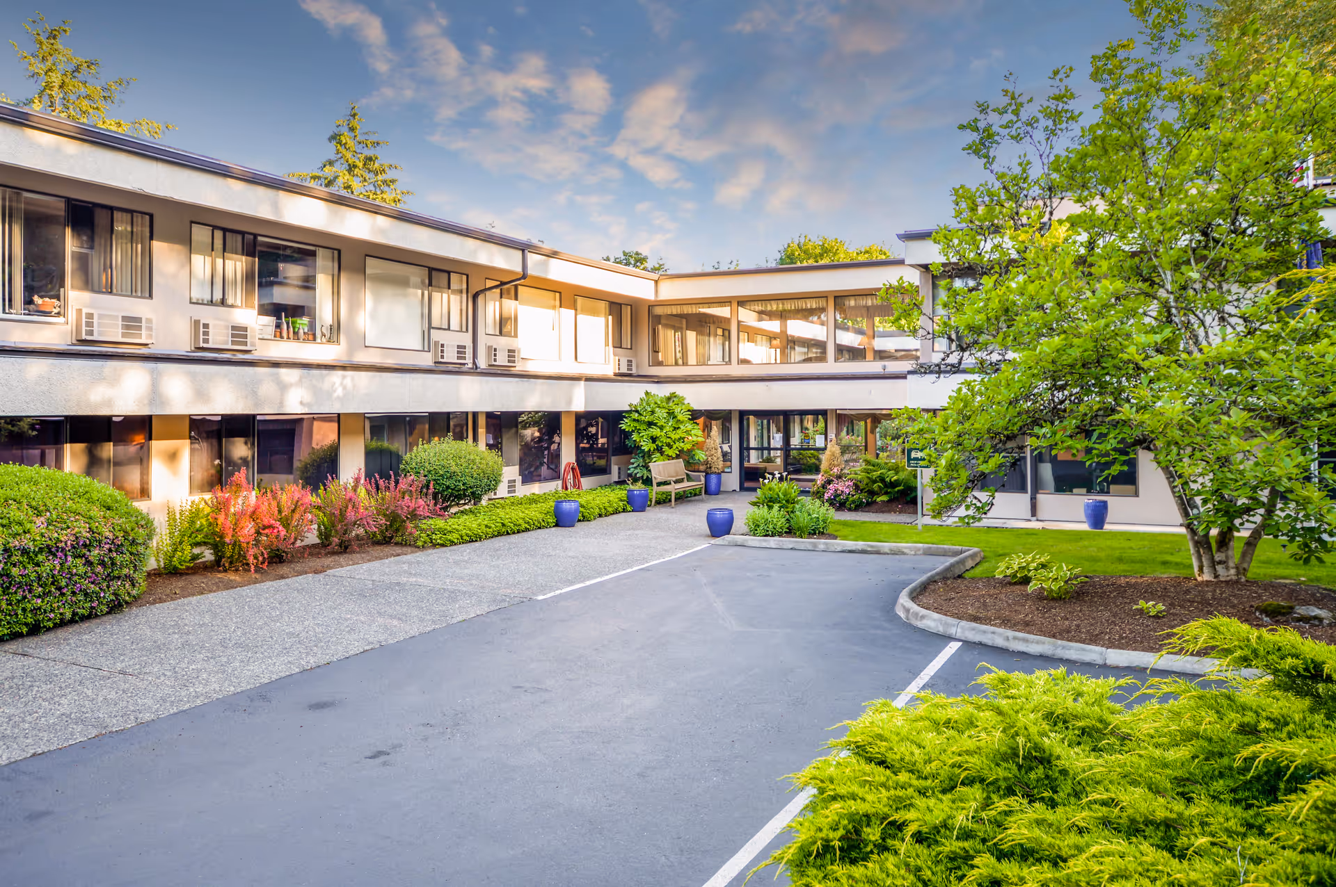 Two-story assisted living building with a landscaped driveway, potted plants, and trees under a partly cloudy sky.