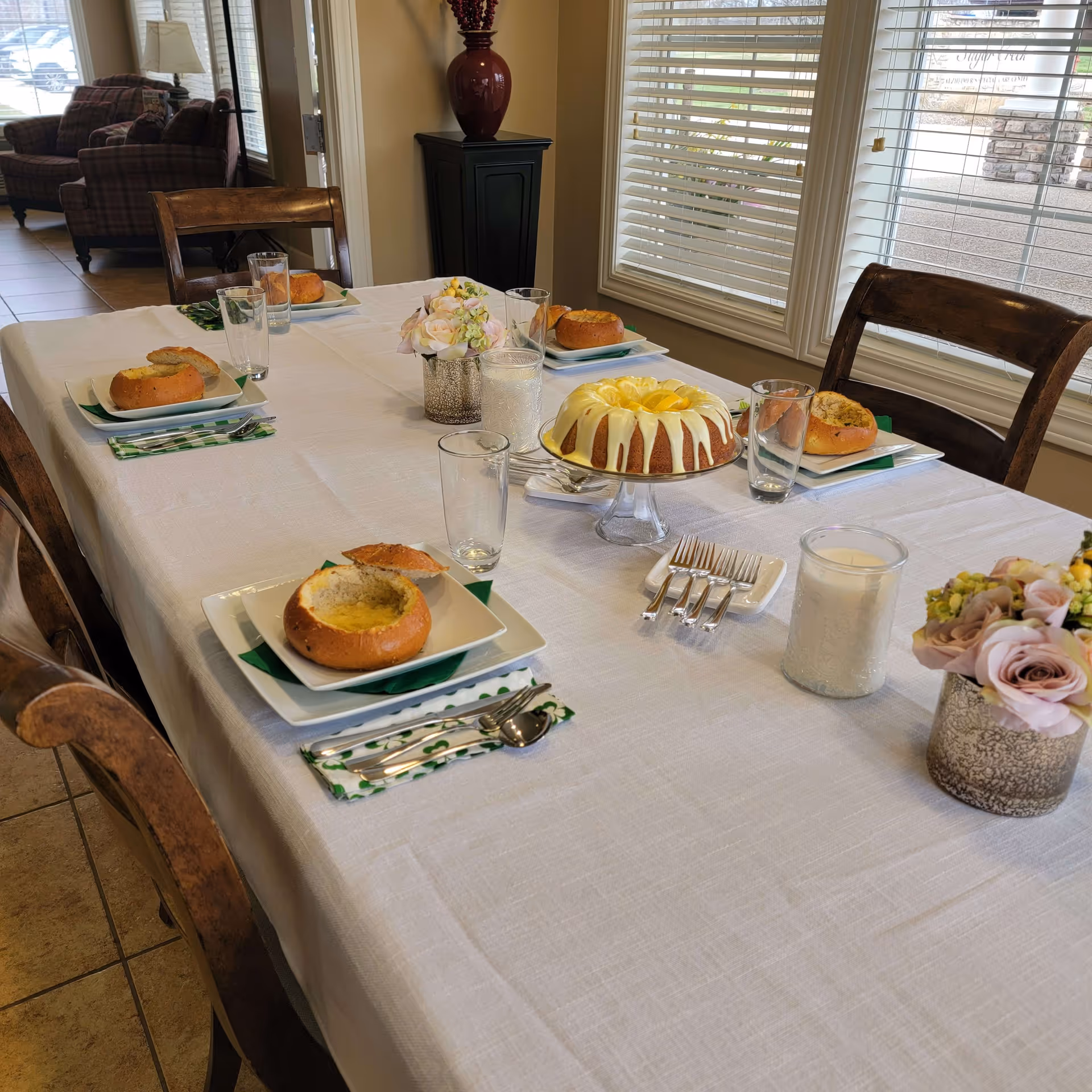 A dining table set for four with white tablecloth, each place setting having a bowl of soup served in a bread bowl, a glass, and utensils on a green and white napkin. In the center of the table is a cake with white icing and yellow decoration on a glass cake stand, a white candle in a glass holder, and a small flower arrangement in a decorative vase. The room has large windows with blinds and wooden chairs around the table. In the background, there is a sitting area with plaid armchairs and a lamp.