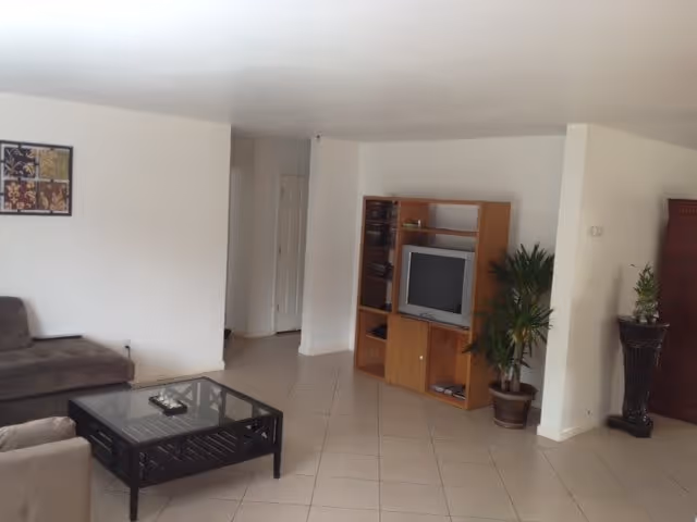 A living room with a brown sectional sofa, a glass coffee table, a wooden entertainment center with a TV, and two potted plants. The floor is tiled, and the walls are white with a framed artwork hanging on one wall.