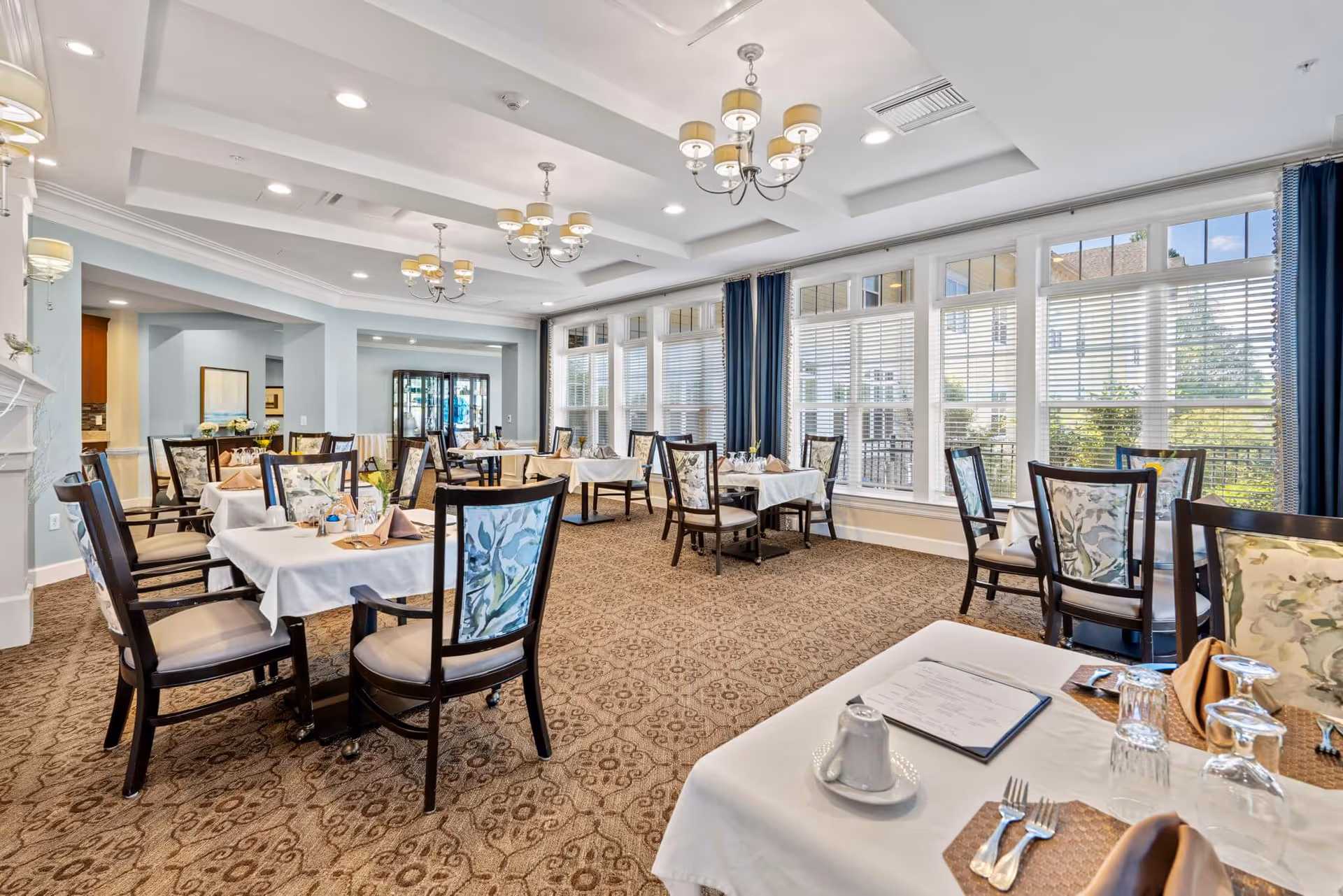 A bright and elegant dining room with multiple tables covered in white tablecloths, set with napkins, glasses, and silverware. The room features large windows with white blinds and blue curtains, patterned carpet, and several chandeliers hanging from a coffered ceiling.