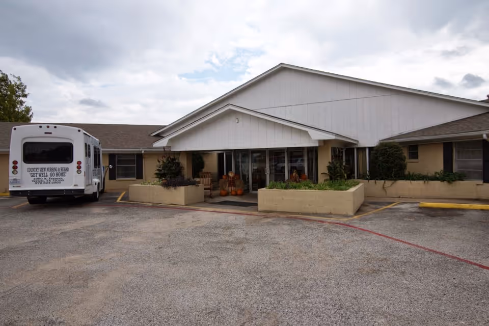 Exterior view of Country View Nursing & Rehabilitation facility showing the main entrance with a covered porch, planters with greenery, and a parked white shuttle bus on the left side. The sky is cloudy.